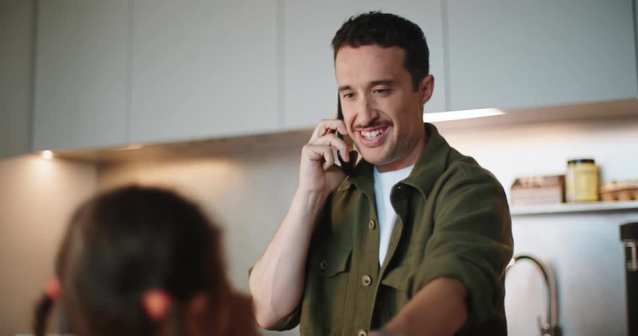 A man is preparing food on a counter while talking on the phone across from a little girl eating a cherry tomato