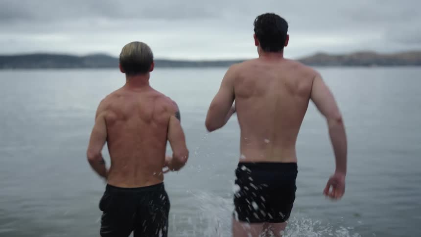Two men are on a beach presumably and dive into the ocean and start swimming