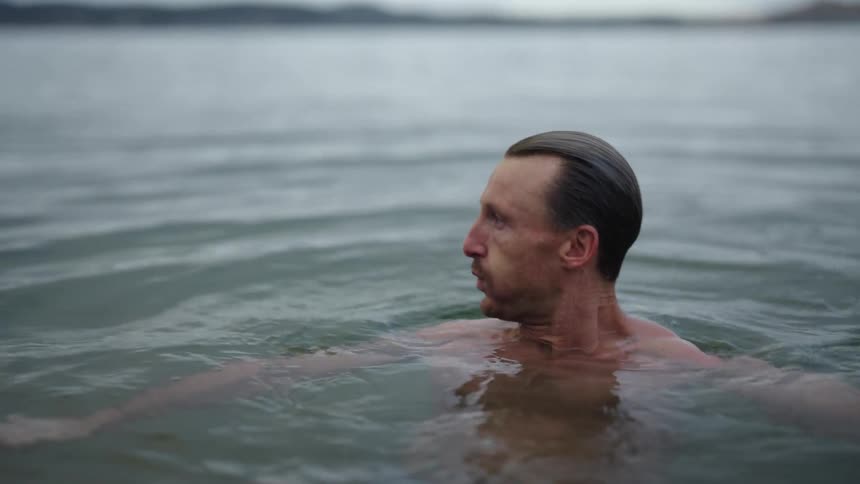 A man is swimming in the lake and he bounces underwater and comes back up and shakes his head