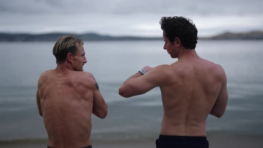 Two man with their bare backs to the camera stretching and then running to the water