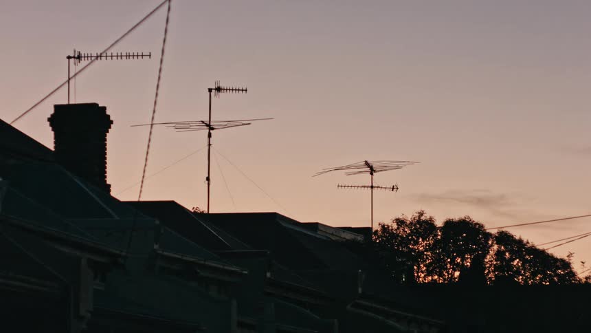 A silhouette of the tops of houses with satellites and power lines atop at dusk