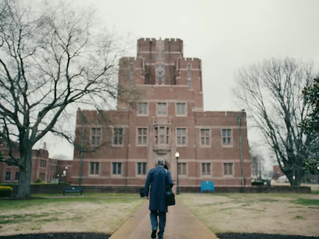 A man in a blue suit carrying a satchel walks up a path to a large brick building