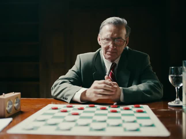 An older man sits down at a table with a board game on it shakes the opponents hand and starts playing
