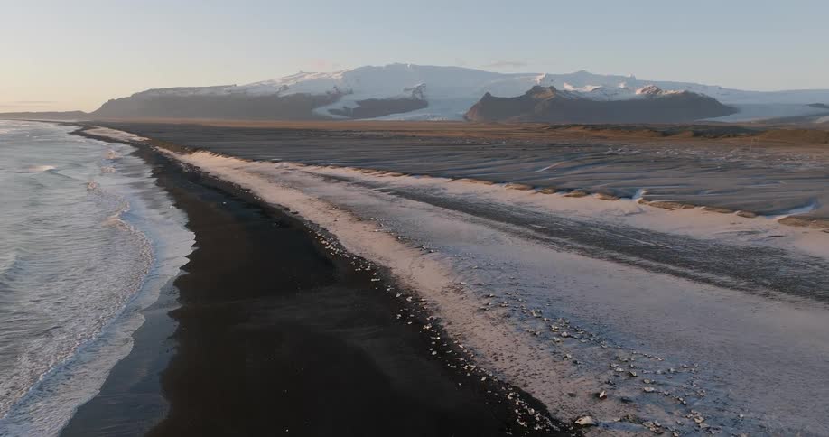 Aerial view of ocean waves gently crashing on a beach near a snow covered mountain range at evening