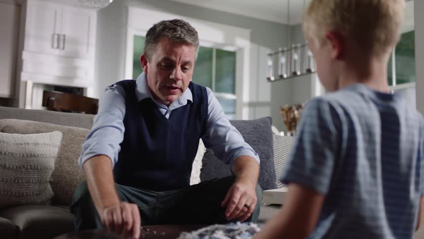 A little boy kneels over a table with pieces of a jigsaw puzzle as his grandfather holds a piece and explains it to the boy