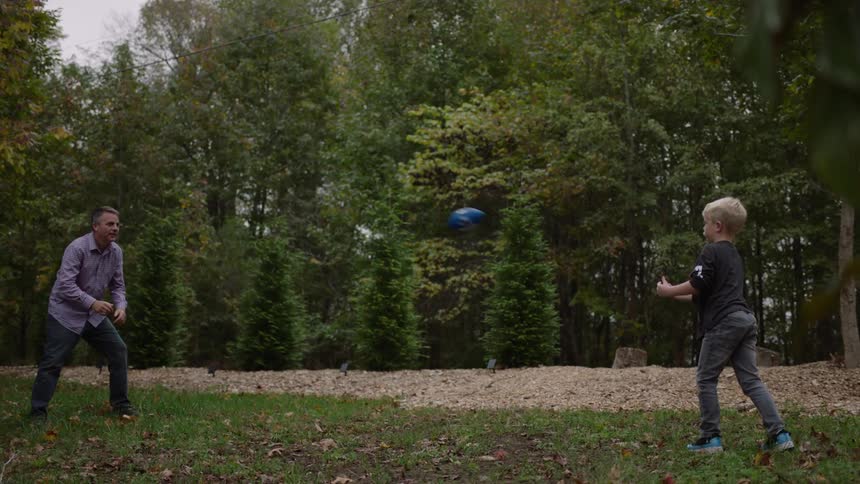 A young boy throws a football to his grandfather as they play outside near the woods