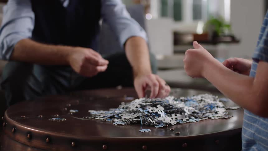 Puzzle pieces piled up on a table while a child and older man pick through the pieces