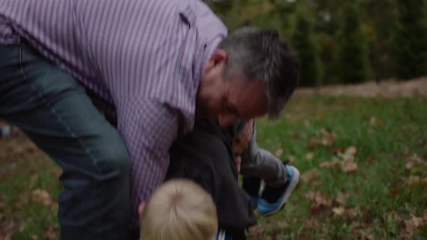 A man is playing lifting his young son up who is facing down laid across his arms he drops him down towards the grass in a wooded area then picks him back up and sets him on his feet smiling the whole time
