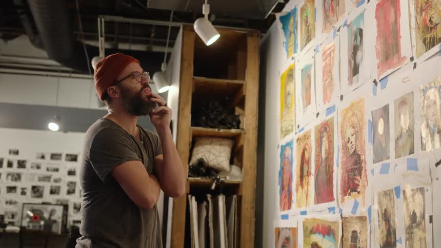 A man standing in front of a wall of paintings with his hands on his face looking contemplatively