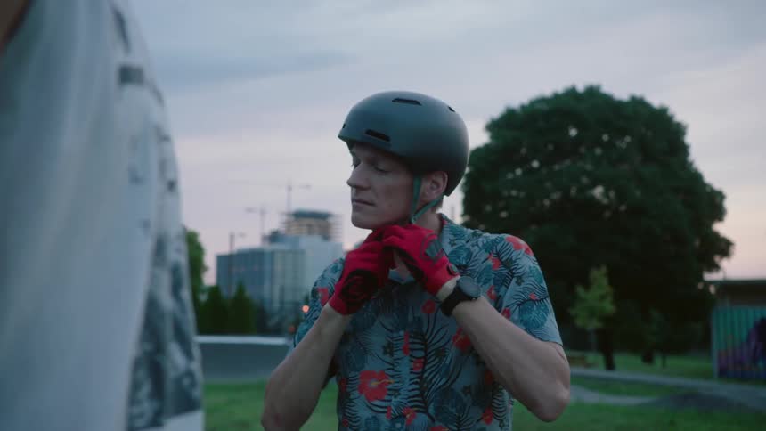 Two men stand next to each other as one puts a helmet on wearing red gloves then they walk away together