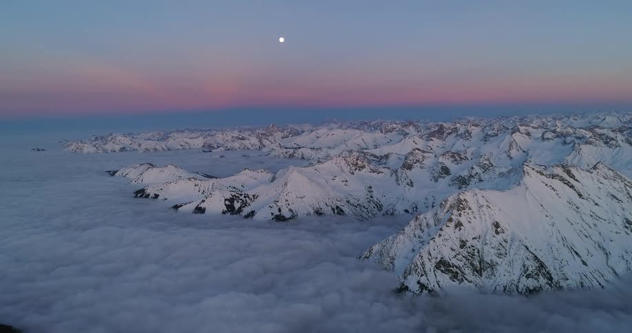 Aerial view of the full moon over the snow covered slopes of a mountain range surrounded by clouds at evening