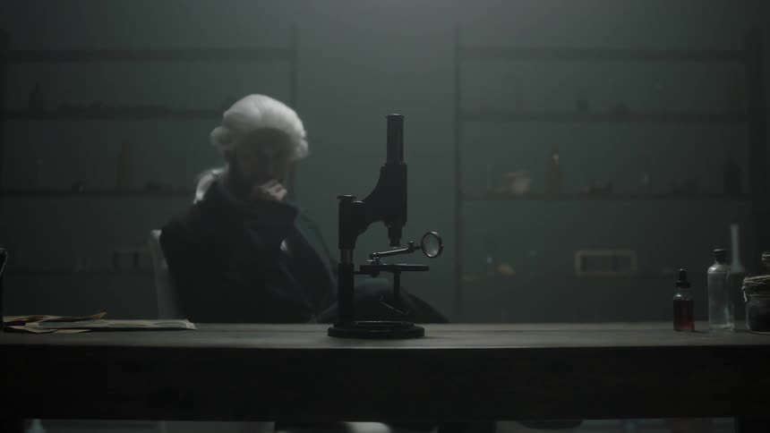 A man in a white wig sits at a desk with a microscope