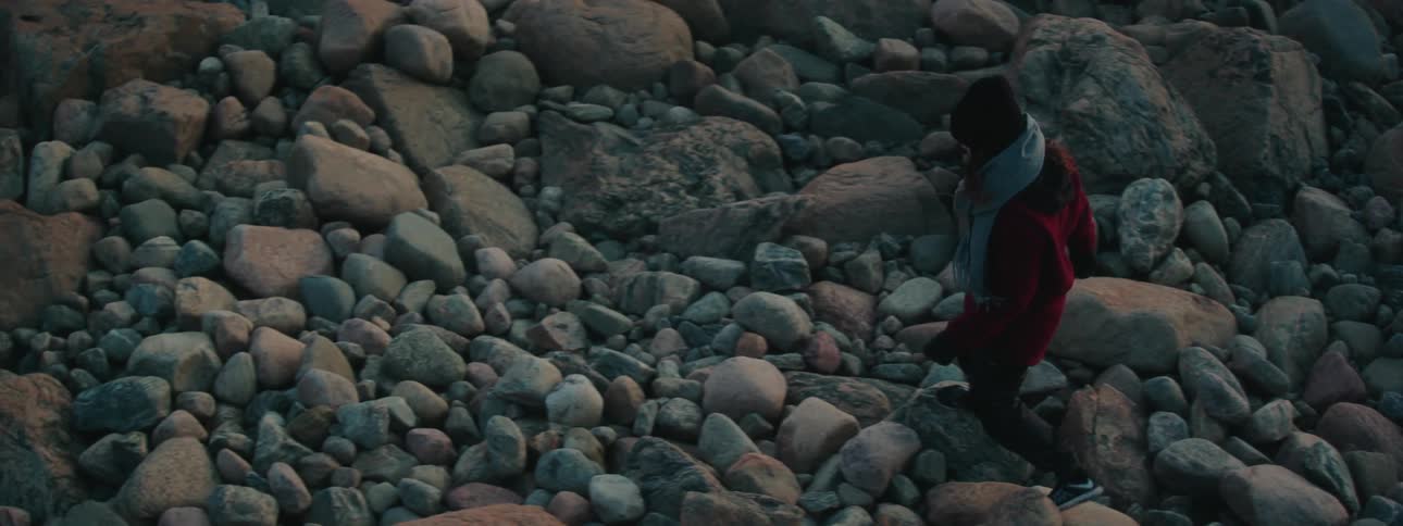 Woman with hat scarf coat and pants walks up on stony shore