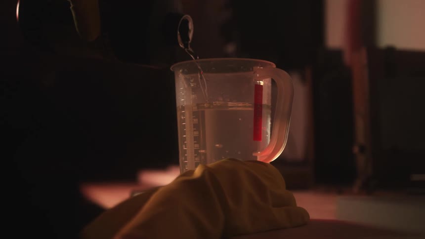 Person wearing gloves pours from bottle into a beaker in dimly lit lab