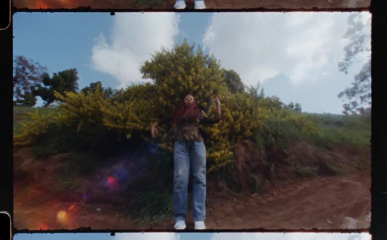 A woman moving her arms and body in front of a large bush on the side of a dirt path