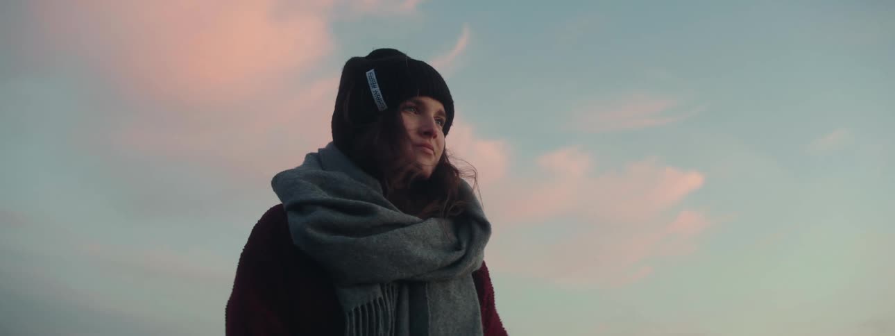A woman with a scarf and hat on stands looking out into the distance
