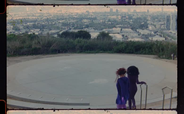 Two friends climb down stairs to flat circular area where they dance with city scape in the background