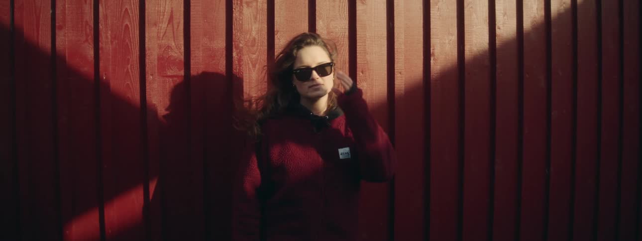 A teenage girl stands against a red wooden fence or house while the wind blows her hair
