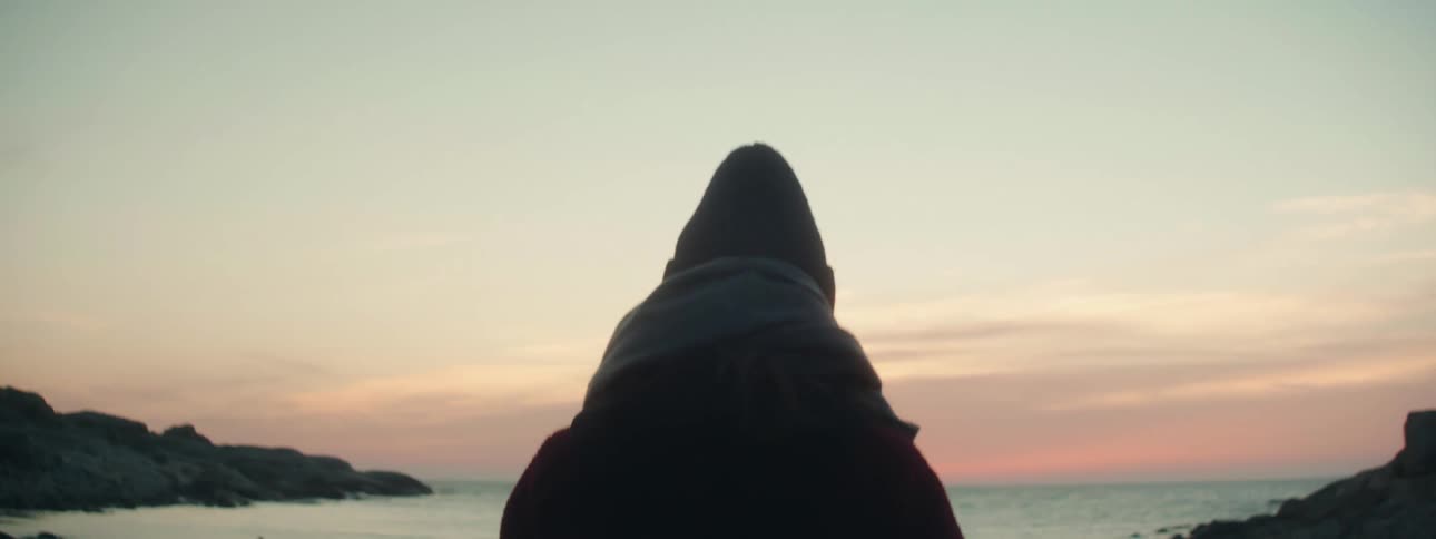Woman with hat scarf and coat walks to the sea at sunset on the pebble beach