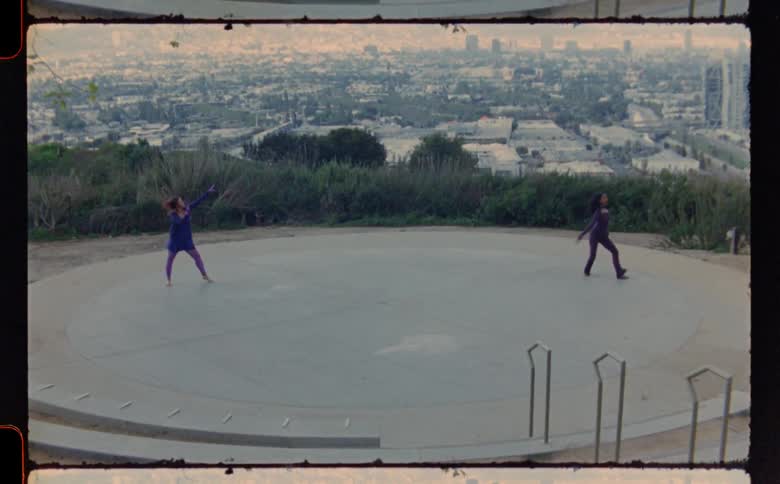 Two women dancing on a concrete circle with a city in the background
