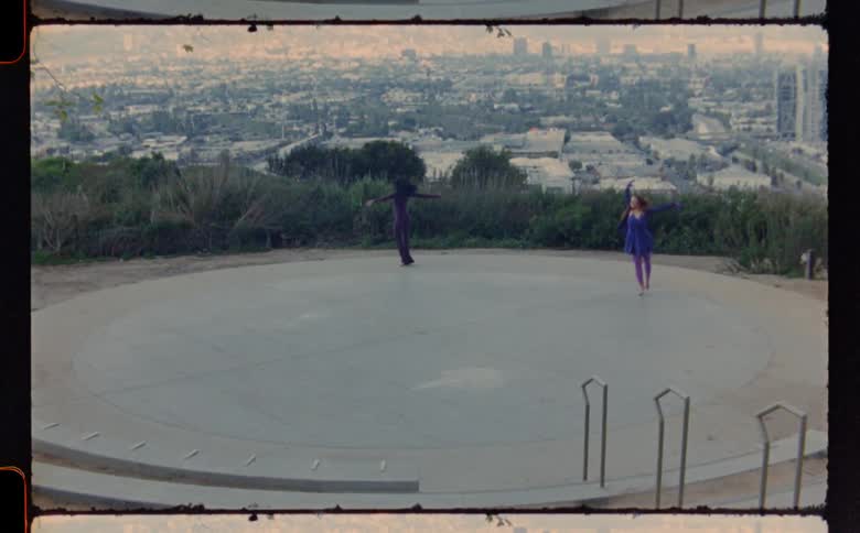 Two women dancing and twirling on a concrete area with a city in the background