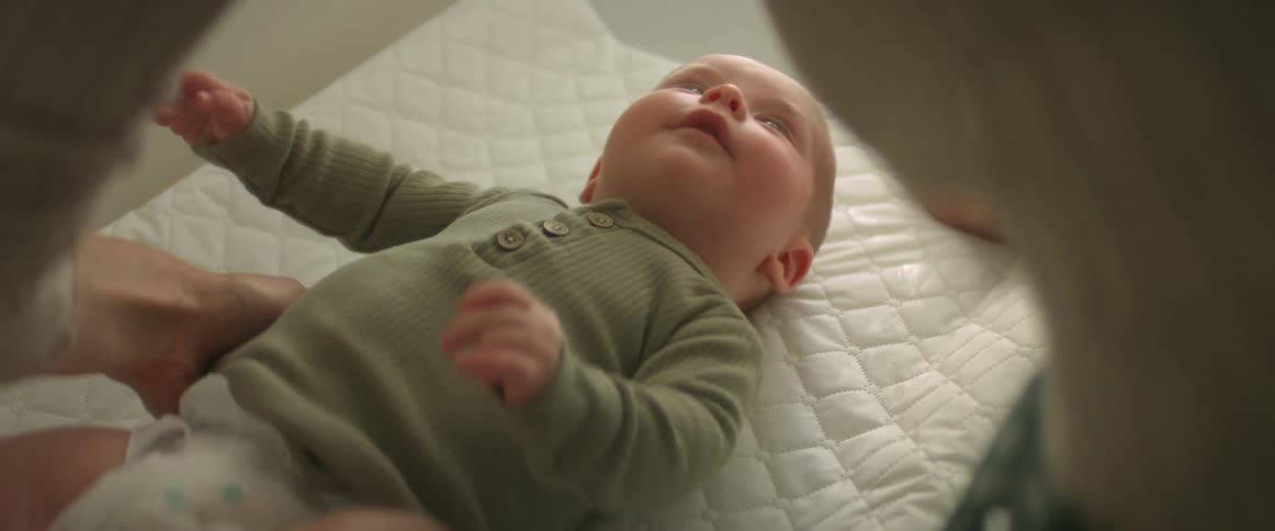 A happy newborn baby lays on a changing table as an adult female braces him with her hand while standing over him