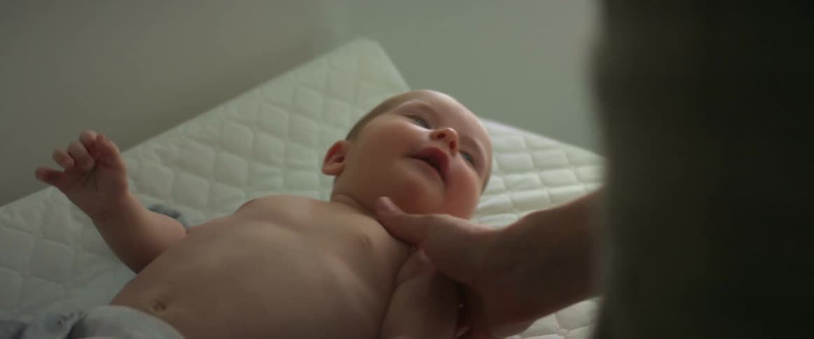 A baby without a shirt on lays on a changing pad as an adult touches the babies shoulder
