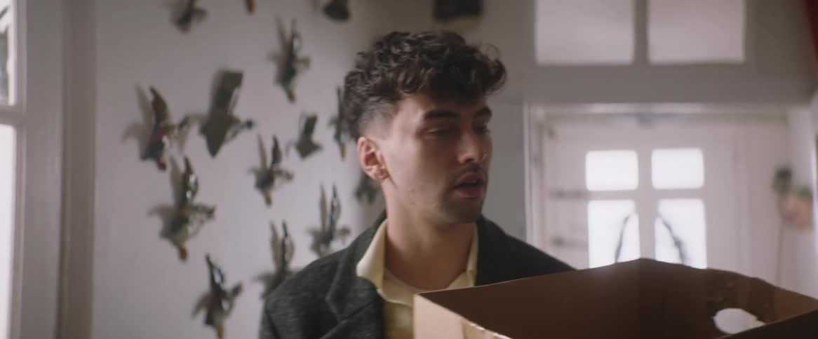 A man carrying a cardboard box opens the door to his home and walks slowly down the hall