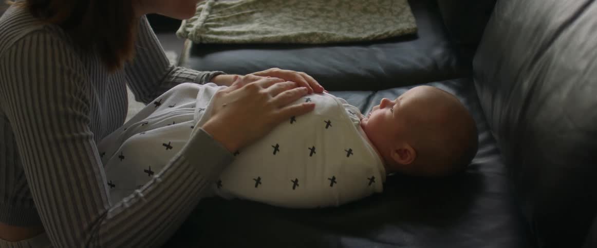 A mother swaddles her infant son in a blanket as he sleeps on the couch