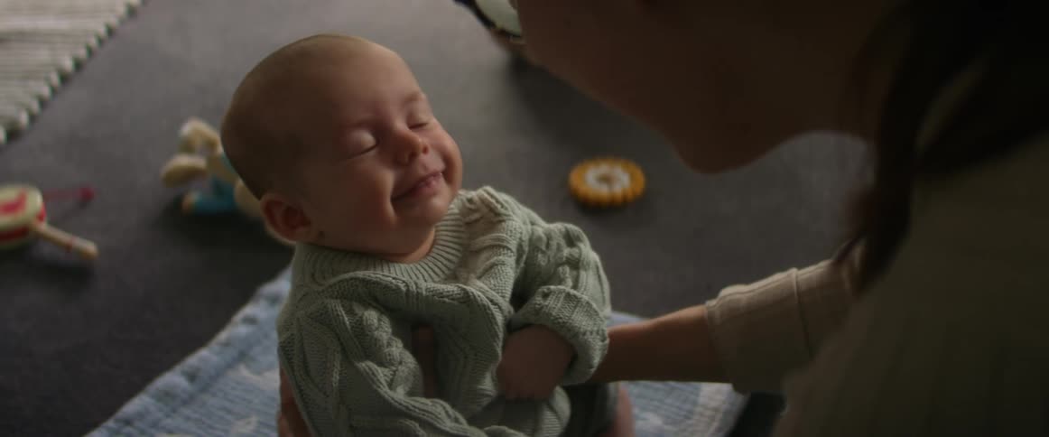 Mother helps baby sit up on blanket on floor near toys
