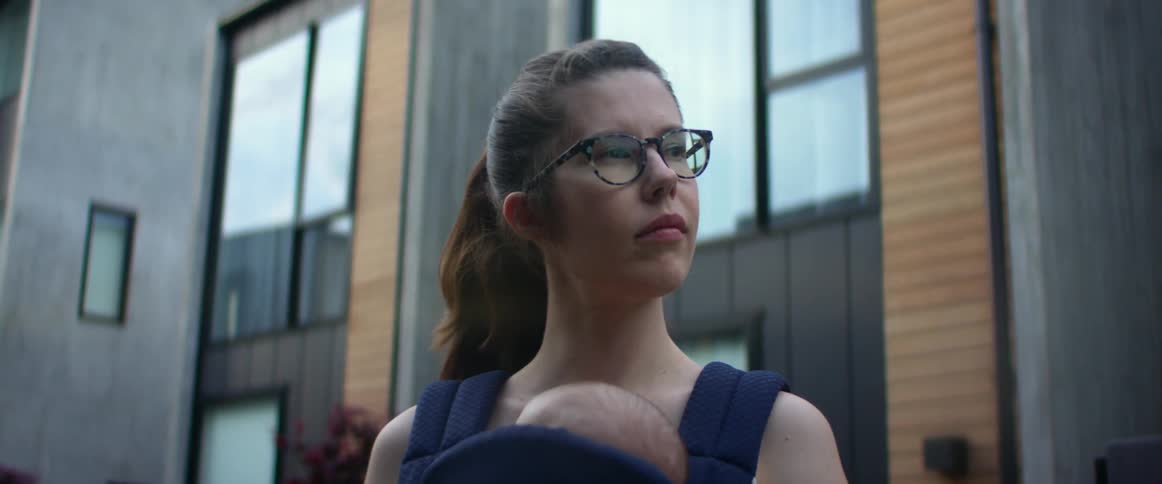 A mother holding her baby in a front carrier looking into the distance in front of a building