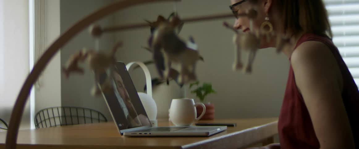 A woman wearing glasses and earrings is chatting with a man on a laptop laughing while sitting at a desk with a coffee cup by her side