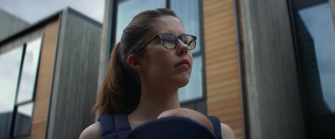 A mother holding her baby in a carrier looking into the distance outside in front of a building