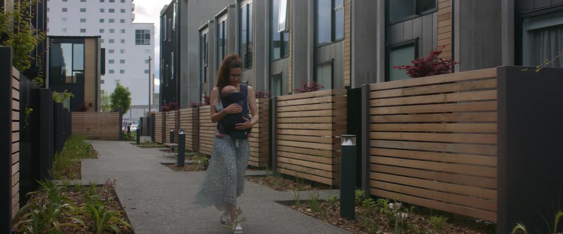A mother walks down a sidewalk with a baby harness strapped to her front with a baby in it