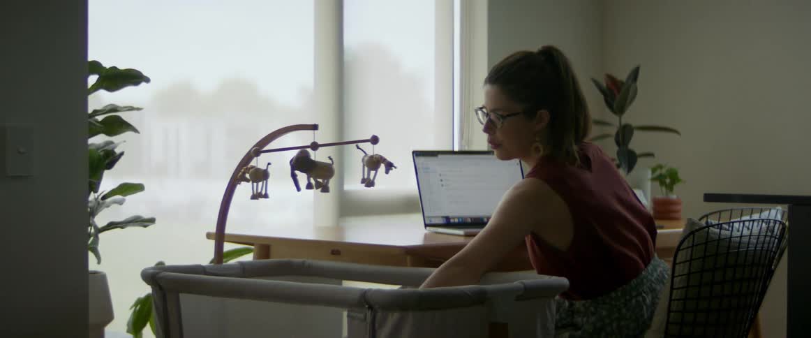 A woman is sitting at a table typing on a laptop computer as she reaches down to comfort her baby sleeping in a playpen next to the table