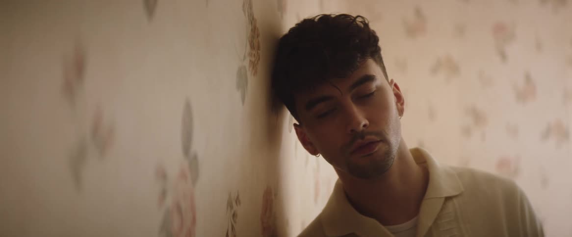 A man is standing leaned over with his shoulder and head on the wall as he looks forward but with his eyes down towards the wall in a worried and sad way