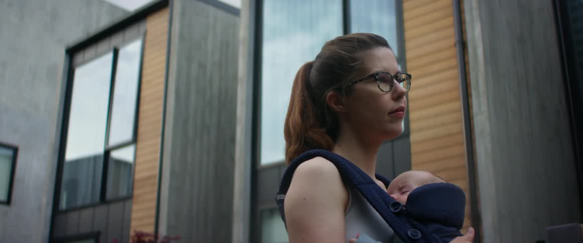 A woman carries a sleeping baby in a front baby sling outside while standing