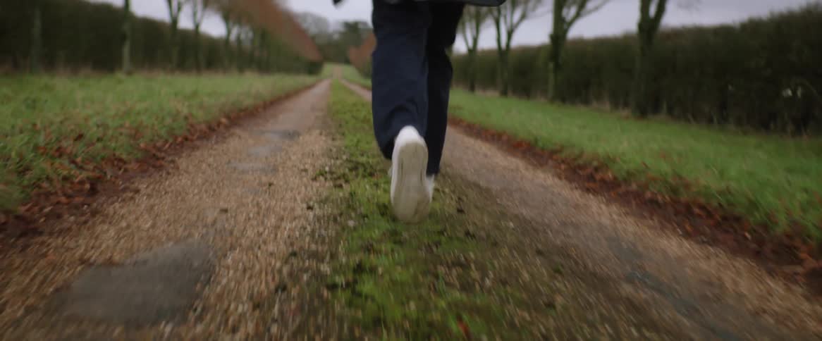 A teenage boy wearing white tennis shoes and a coat is running on a dirt road lined with trees