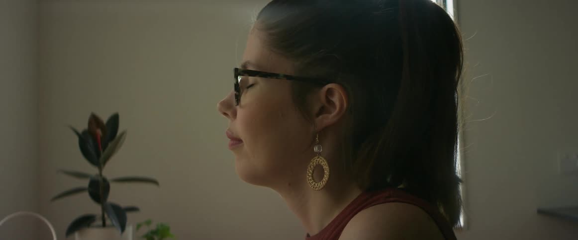 A woman wearing glasses and earrings is standing in aroom with her eyes closes and she smiles and opens her eyes as she looks ahead