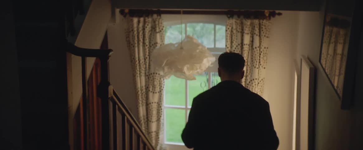 A man runs down the stairs and past a window in a house as if he is hurrying to get somewhere