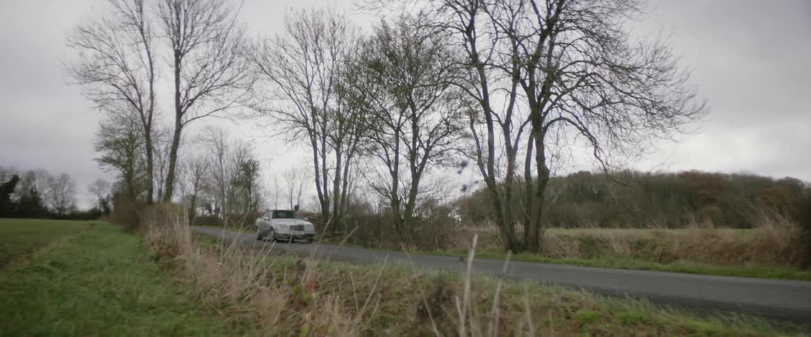 A white automobile travels on a country road surrounded by bare trees