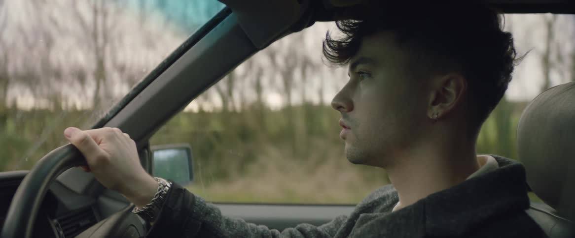 A man is moving his head looking out of the windshield as he drives an automobile on a country road lined with trees