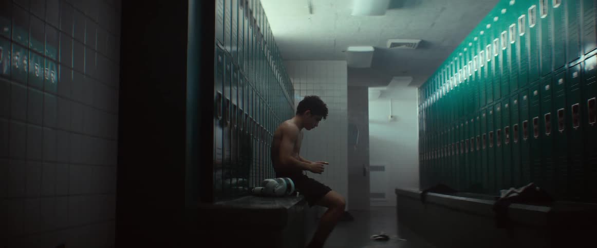 A teenage boy sits in a boys locker room in front of lockers on a bench fiddling with something in his hands