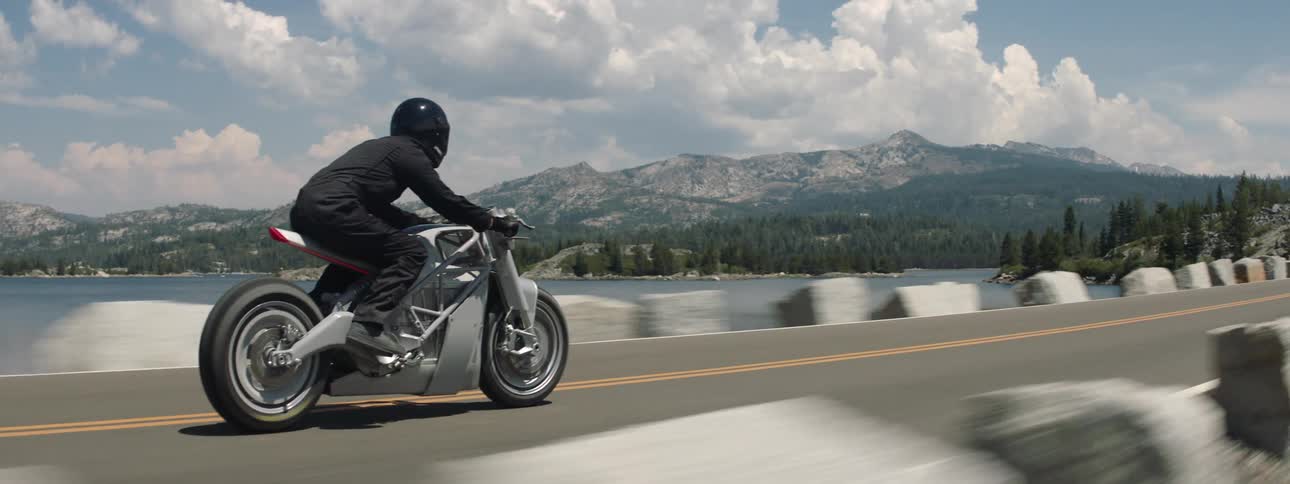 A person wearing black clothes and a black helmet ride his motorcycle on a road near water with mountains in the background