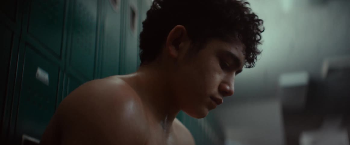 A teenage boy sits in a locker room in front of lockers looking down like he is doing something