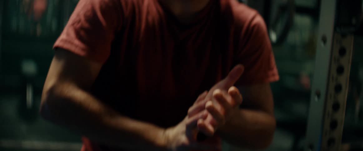 A teenage boy is standing in a gym near weights as he moves his hands and cracks his knuckles