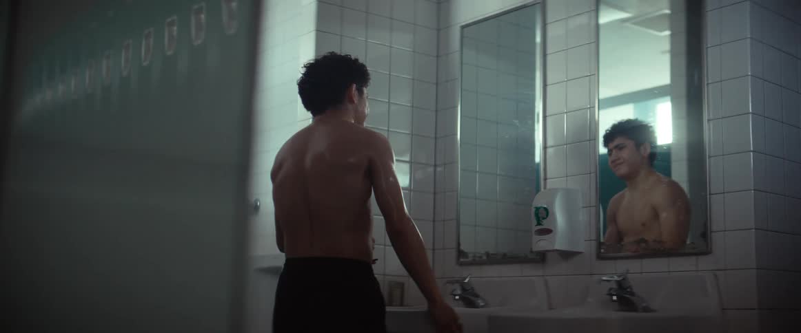 A man without a shirt on walks up to a bathroom mirror and hold his stomach as in pain