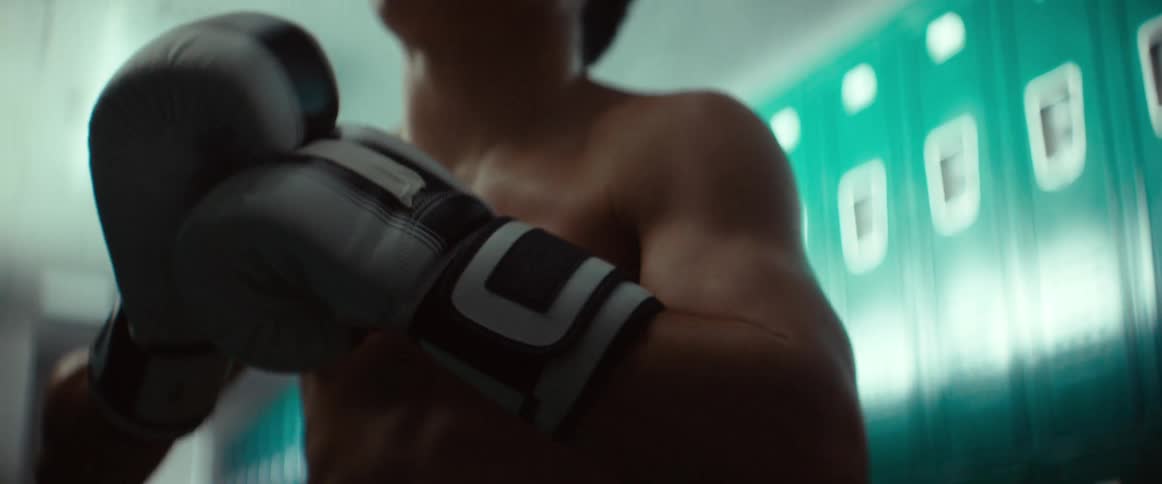A teenage boy wearing boxing gloves is standing in the locker room moving his hands and punching his fists together