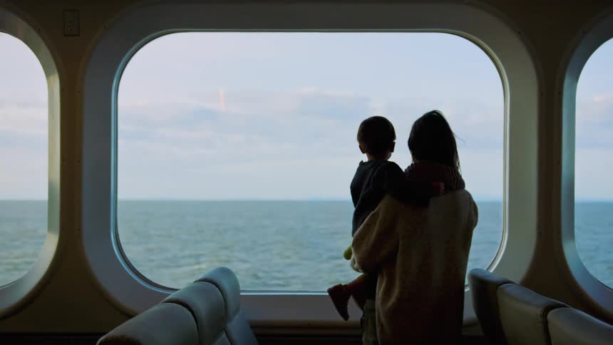 Mother holds son as she stands near ship window showing ocean
