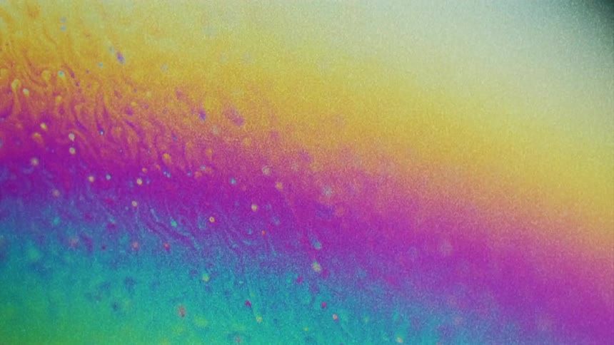 A fluorescent rainbow of colors move as they blend and small particles float over the surface colors as they stretch